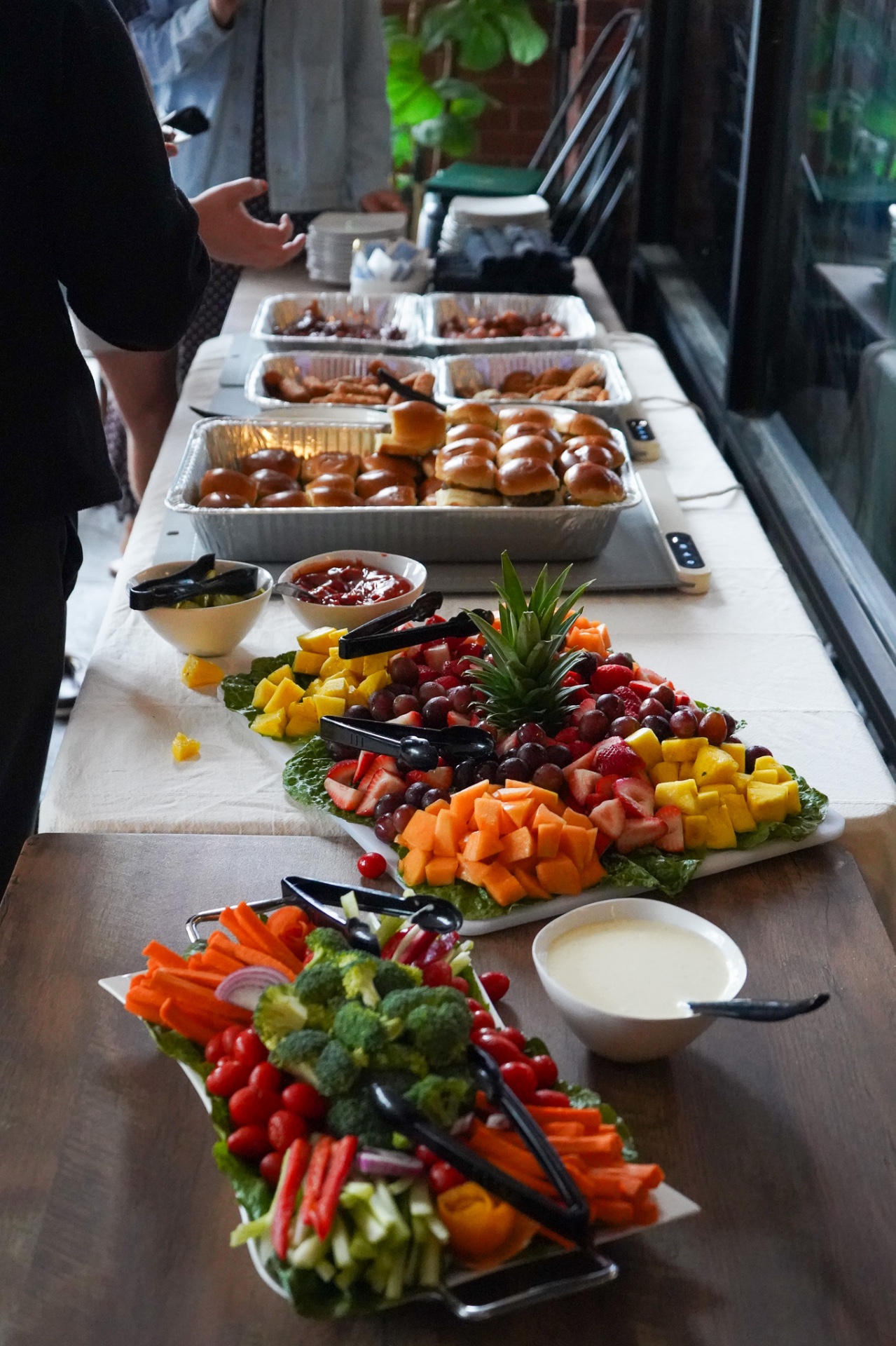 Buffet line with foil trays of sliders, wings and fried appetizers on warming trays, plus a fruit platter, vegetable tray and bowls of dip.