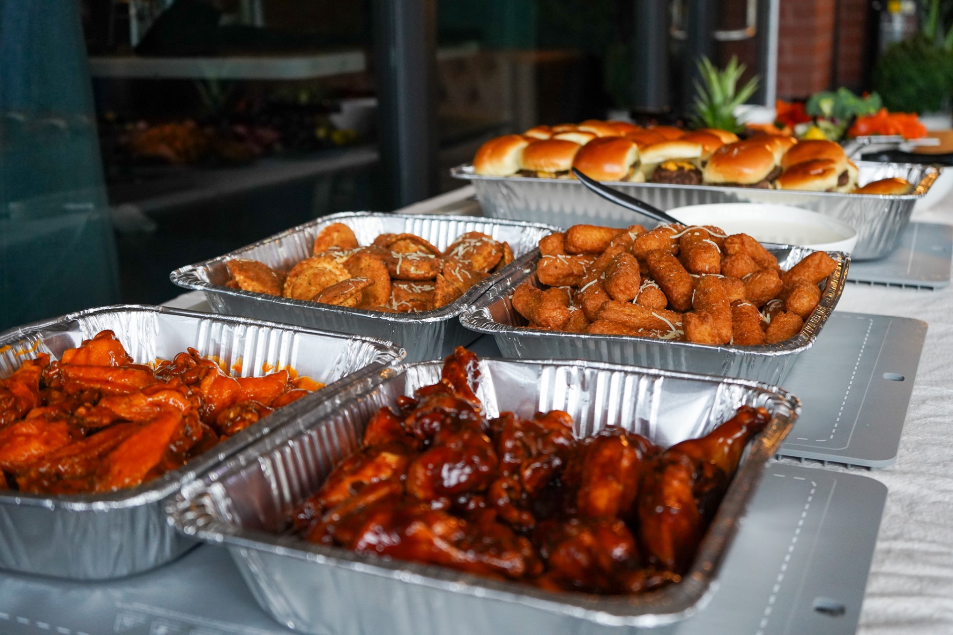 Table lined with foil trays of buffalo wings, barbecue wings, toasted ravioli, cheese sticks and sliders with a bowl of white dipping sauce.