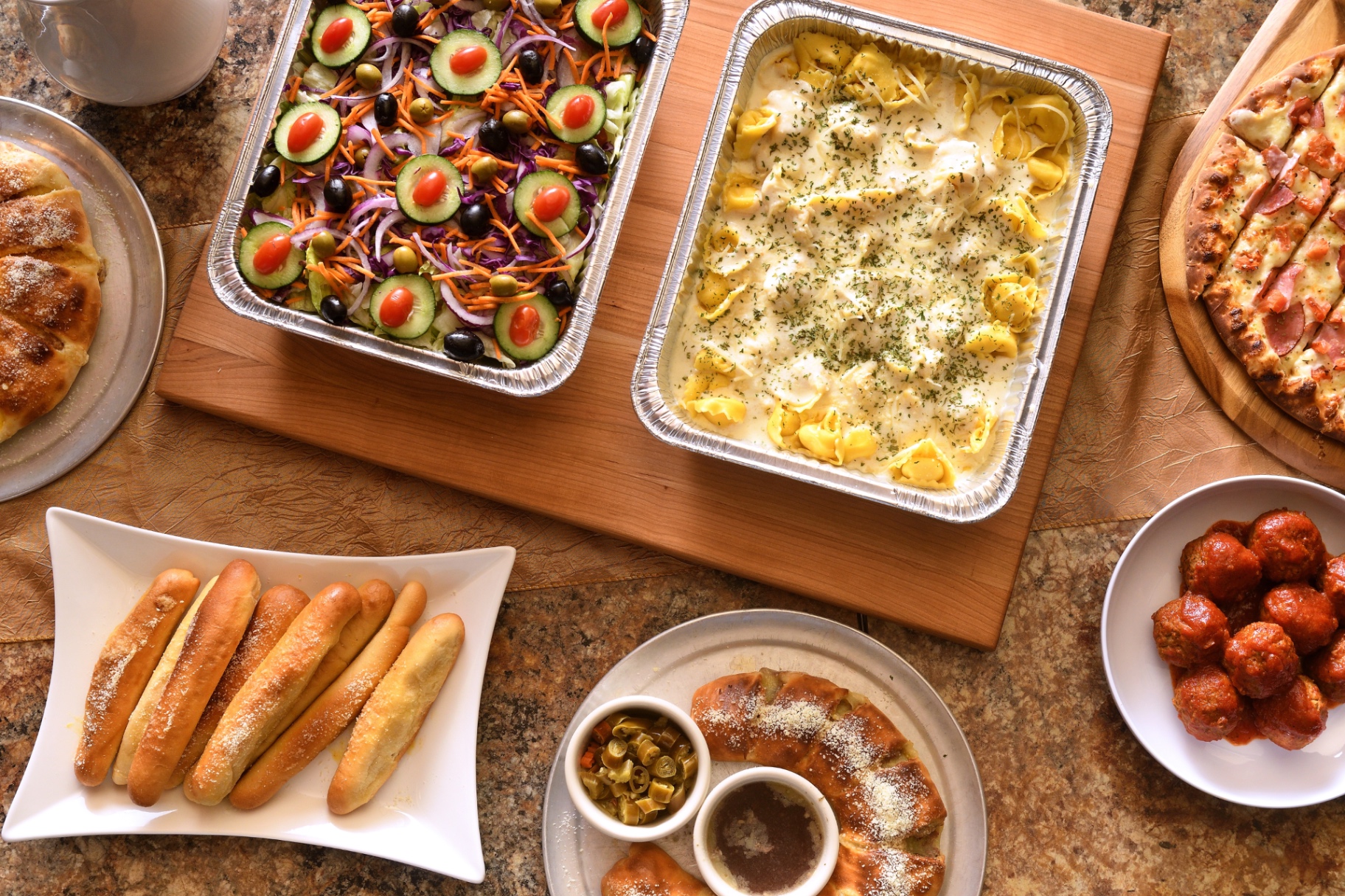 Catering spread with a garden salad tray, a tray of tortellini in cream sauce, pizzas, breadsticks, a bowl of meatballs and dipping sauces.